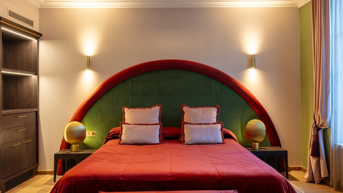 Grande tête de lit verte avec bordure rouge, lit à baldaquin avec couverture rouge et coussins blancs, deux tables de chevet avec lampes sphériques dorées, armoire sombre à gauche et fenêtre avec rideaux à droite