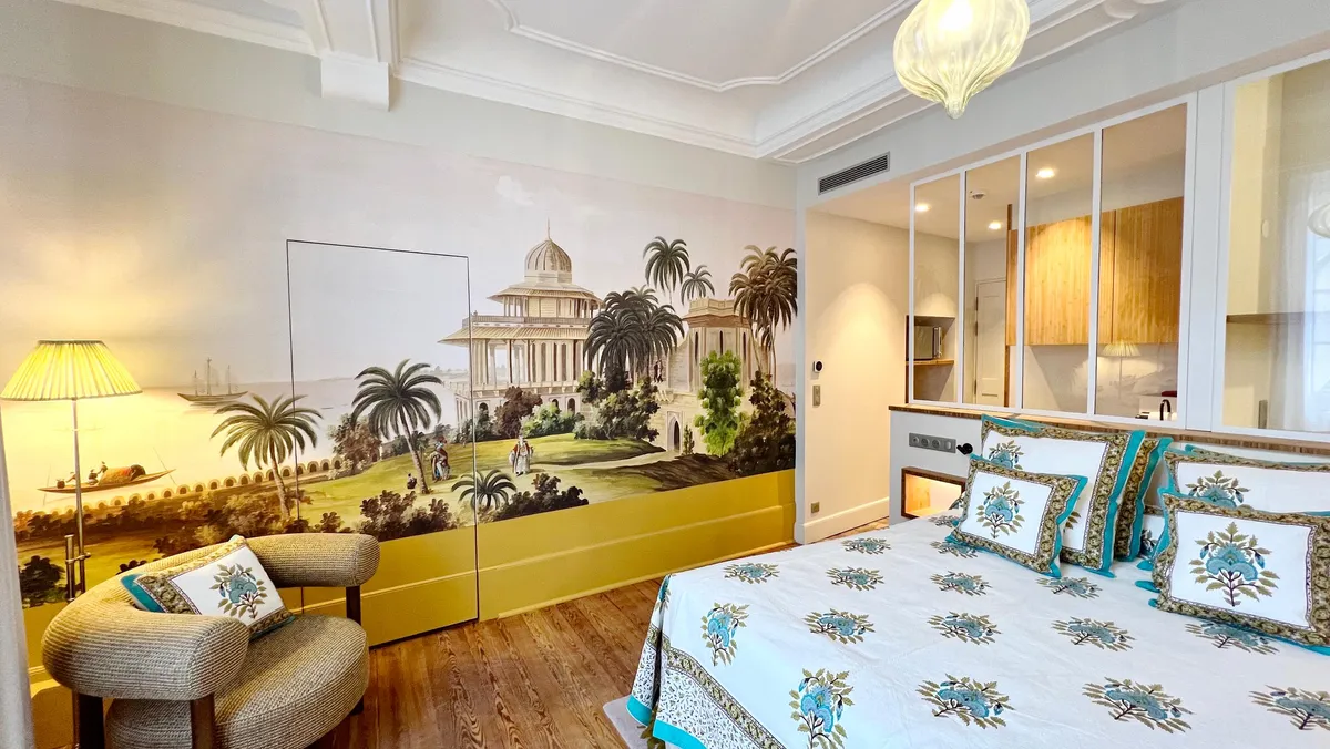Chambre avec un lit à motifs floraux, un fauteuil et une fresque murale représentant un paysage avec des palmiers et un bâtiment classique