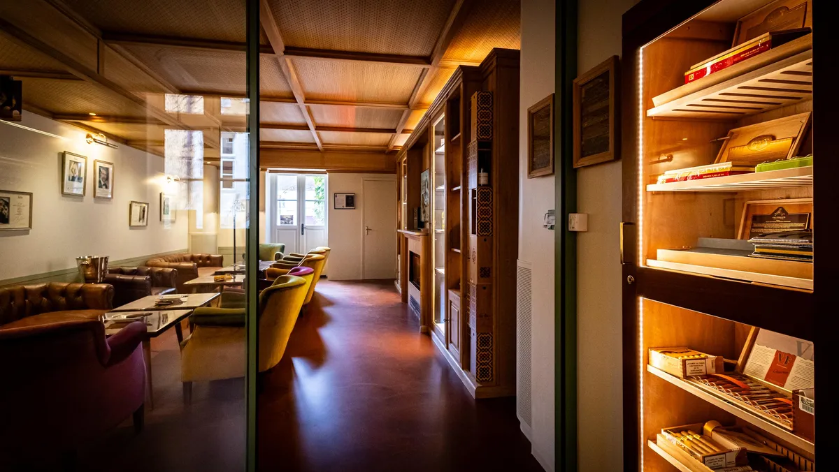 Un couloir avec une porte vitrée menant à une pièce contenant des canapés et des tables, et une étagère remplie de boîtes de cigares