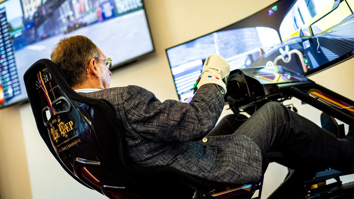 Un homme assis dans un siège de simulation de course, utilisant un volant et des pédales devant deux écrans affichant un jeu de course