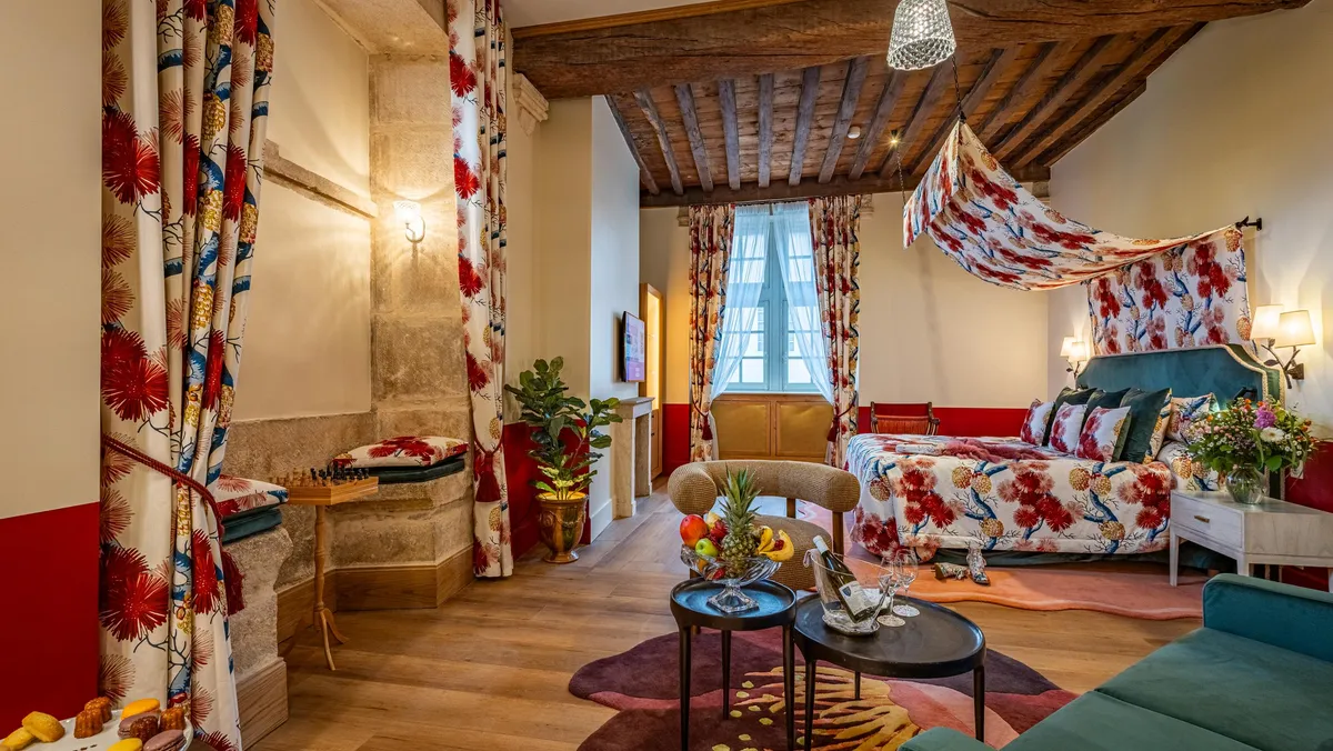 Chambre avec lit à baldaquin, canapés, tables basses et décor floral, sous un plafond à poutres apparentes