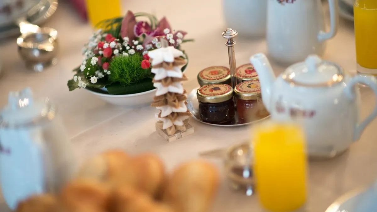 Table dressée avec des verres de jus d'orange, une théière, des pots de confiture, des fleurs et des viennoiseries
