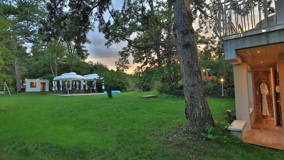 Jardin avec pelouse, arbres et pergola, maison avec terrasse et escalier extérieur, éclairage extérieur allumé au crépuscule