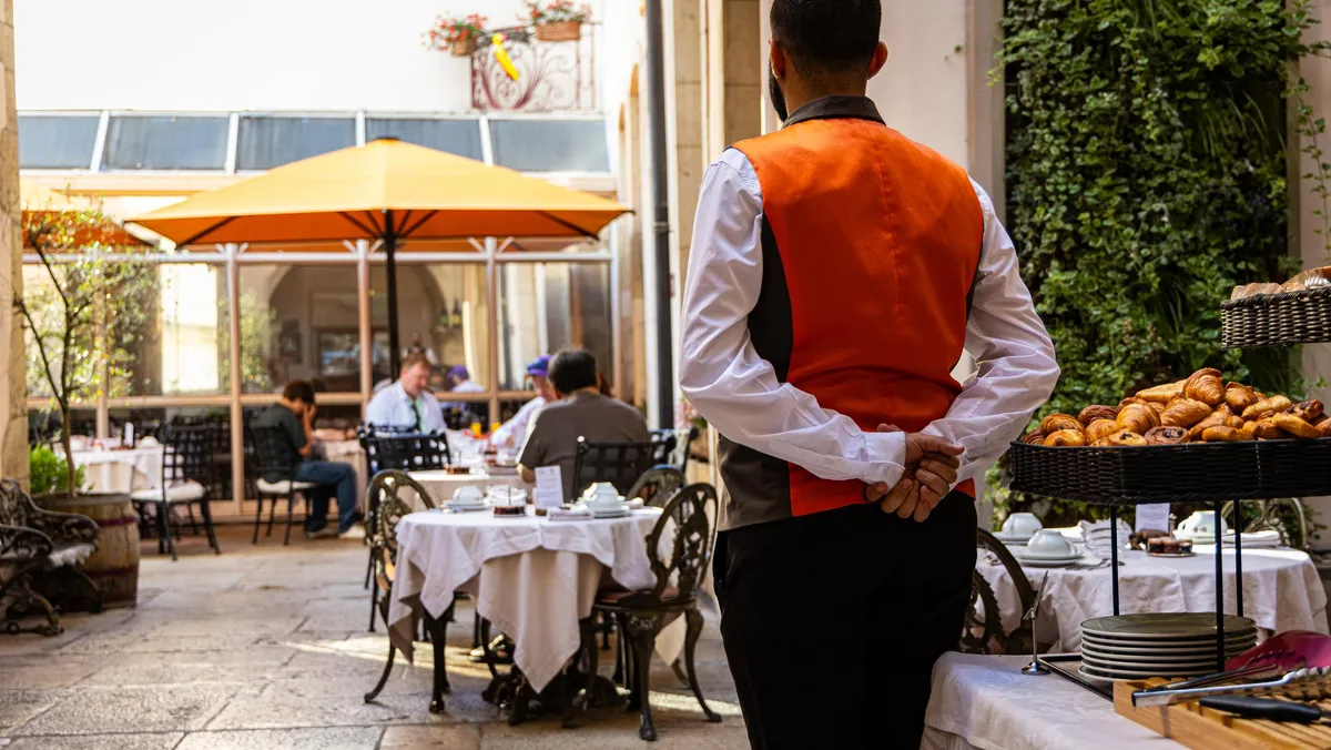 Un serveur en veste orange et chemise blanche se tient dans un restaurant avec des tables dressées et des clients assis sous des parasols orange