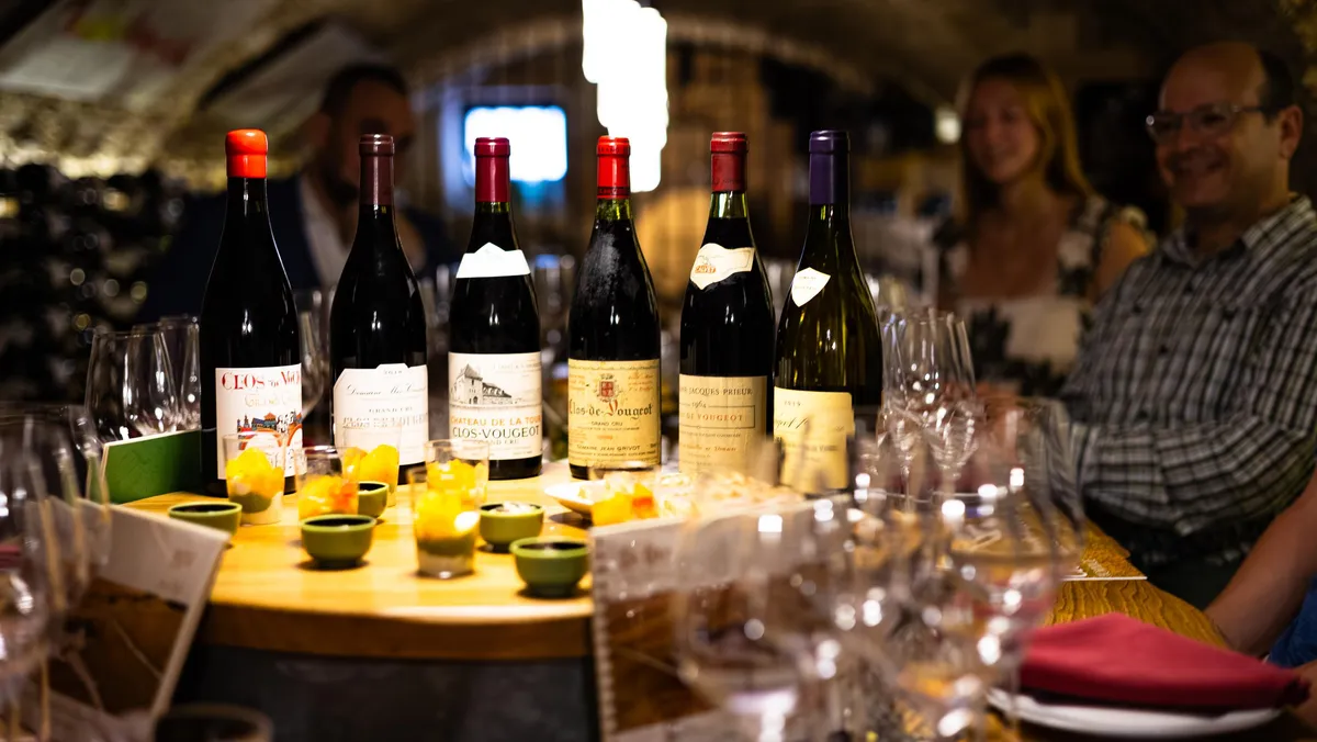 Plusieurs bouteilles de vin et verres à vin sur une table avec des petits bols de nourriture, des personnes assises autour