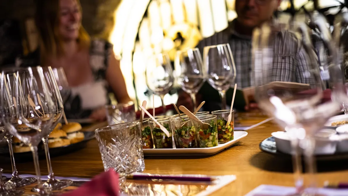 Une table avec des verres à vin, des petits plats contenant des amuse-bouches et des personnes en arrière-plan dans un cadre éclairé