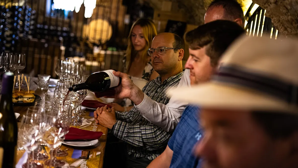 Un homme verse du vin dans des verres à pied sur un comptoir, entouré de plusieurs personnes dans un cadre intérieur éclairé