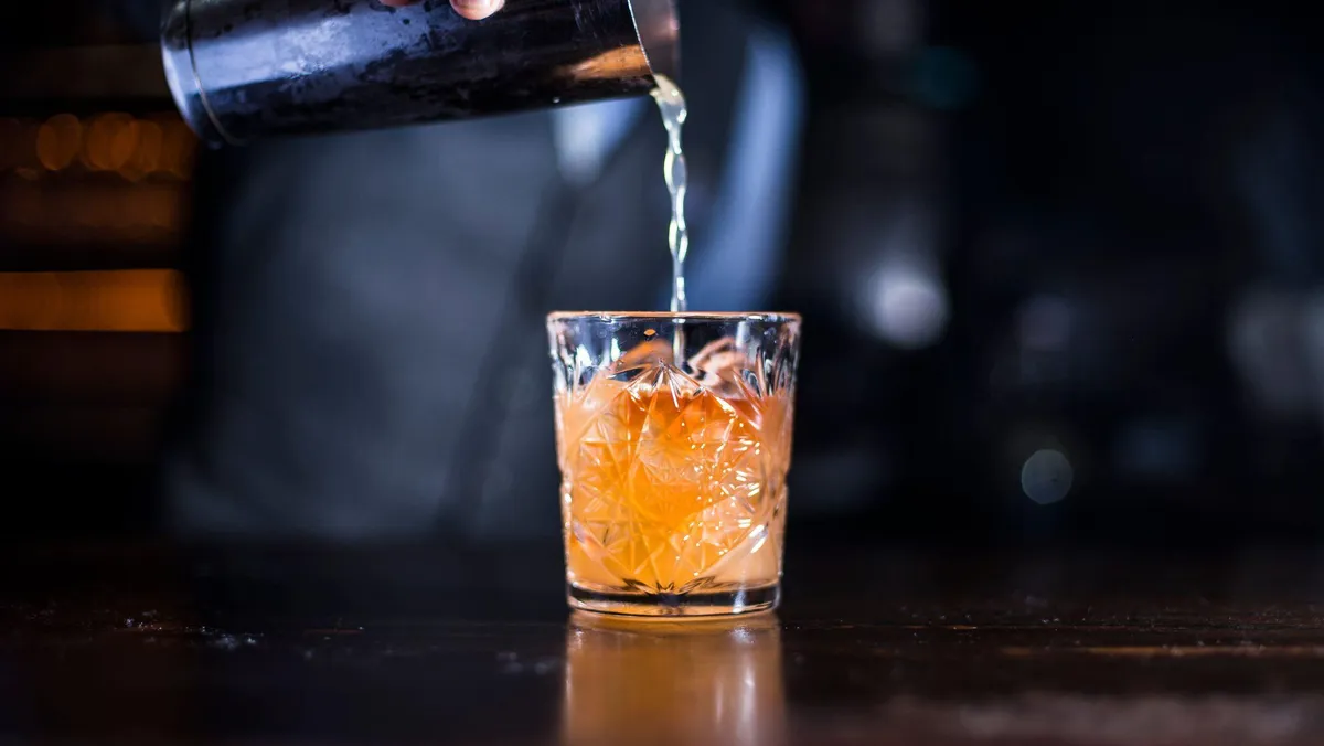 A shaker pours a liquid into a glass containing a large ice cube.