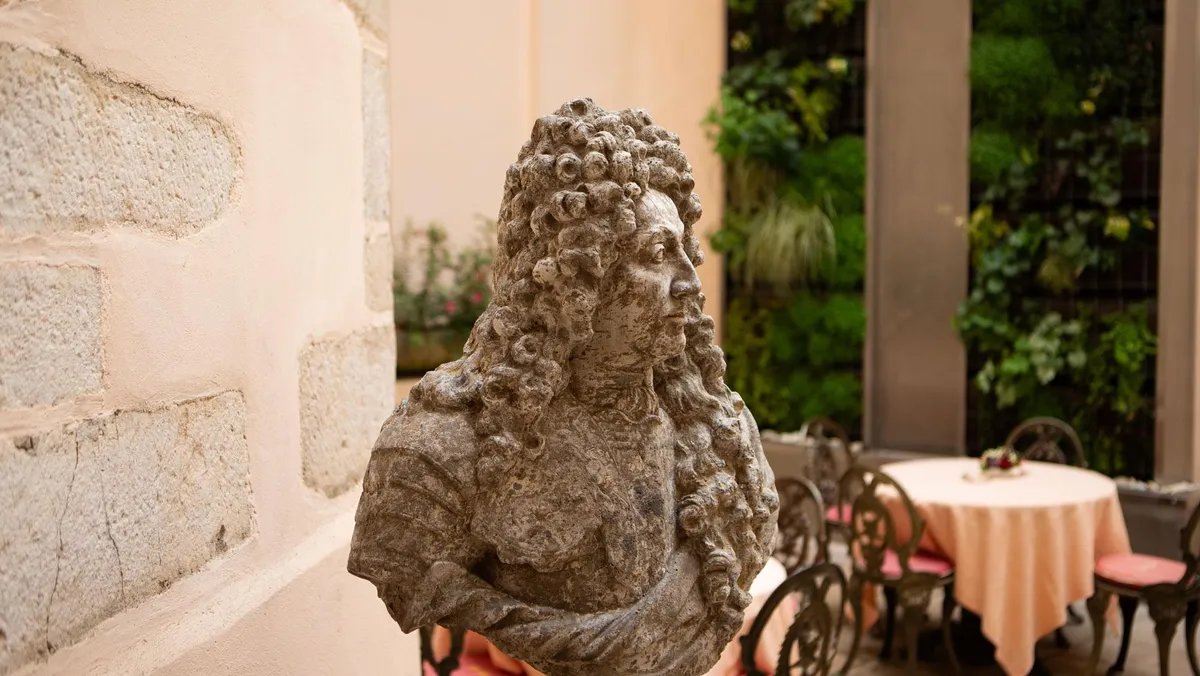 Buste sculpté d'un homme à perruque posé sur un socle, devant un mur de pierre et une table couverte d'une nappe