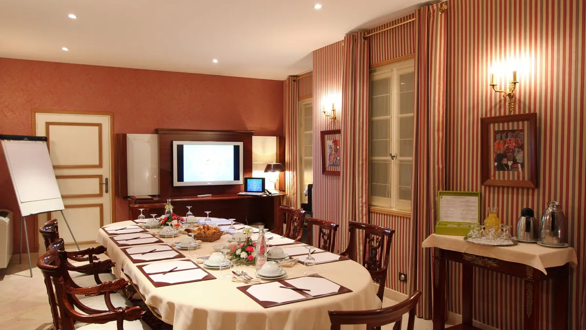 Salle de réunion avec une table rectangulaire couverte d'une nappe blanche, des chaises en bois, un écran, un tableau blanc et une table de service avec des boissons
