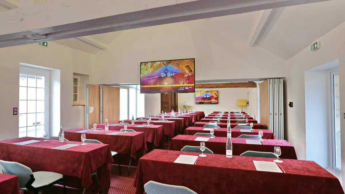 Salle de réunion avec des tables recouvertes de nappes rouges, des verres et des bouteilles d'eau, un écran allumé au fond
