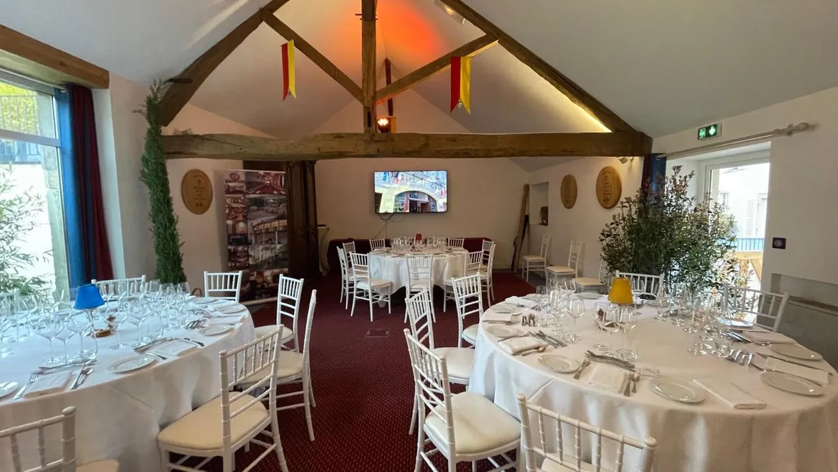Salle de réception avec tables rondes couvertes de nappes blanches, chaises blanches et décorations florales, sous un plafond à poutres apparentes
