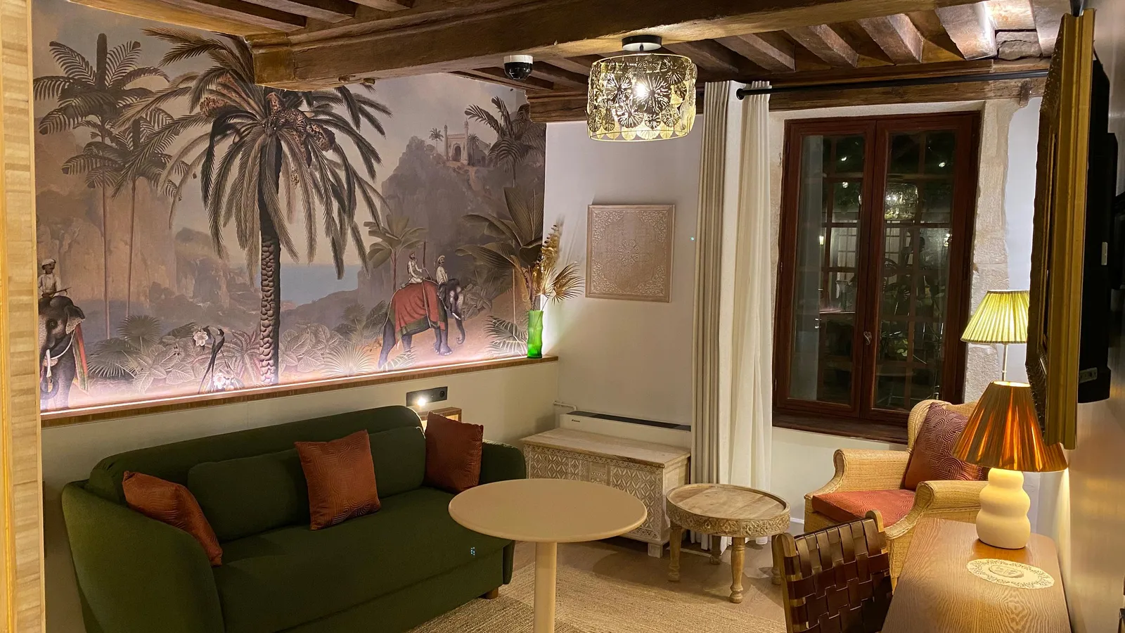 Salon avec canapé vert, fauteuils et tables basses, décoré de coussins et d'une grande fresque murale représentant des palmiers et des cavaliers sur des éléphants