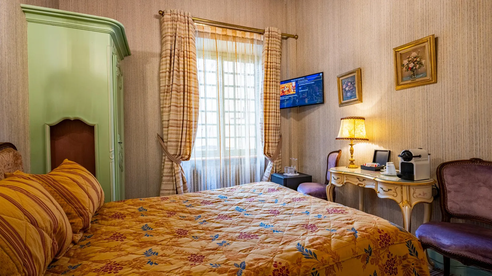 Bedroom with a four-poster bed featuring floral patterns, a window with curtains, a television set, and a bedside table with a lamp.