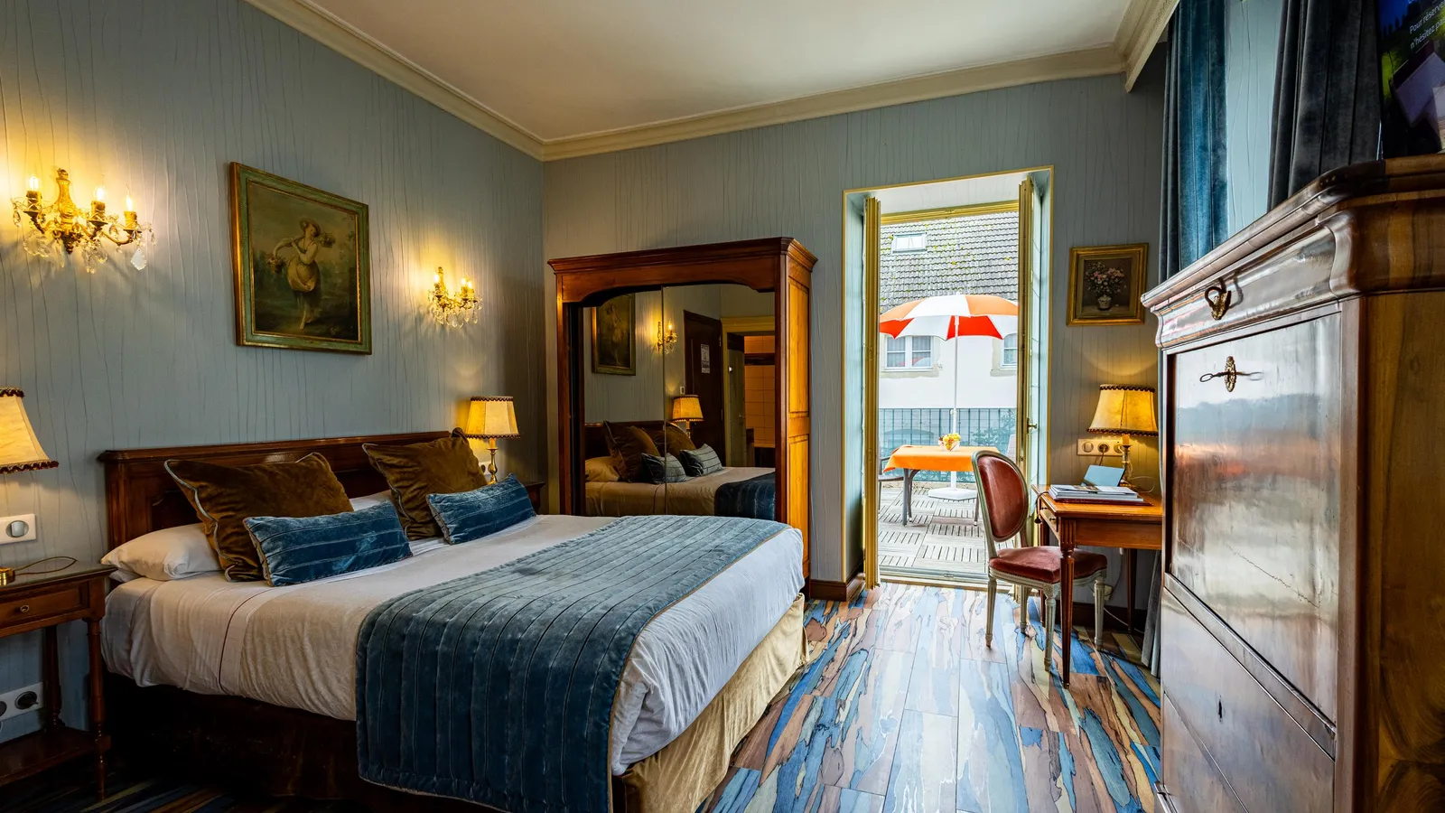 Chambre avec un lit double, une armoire, une porte ouverte sur un balcon avec une table et une chaise, et des lampes allumées