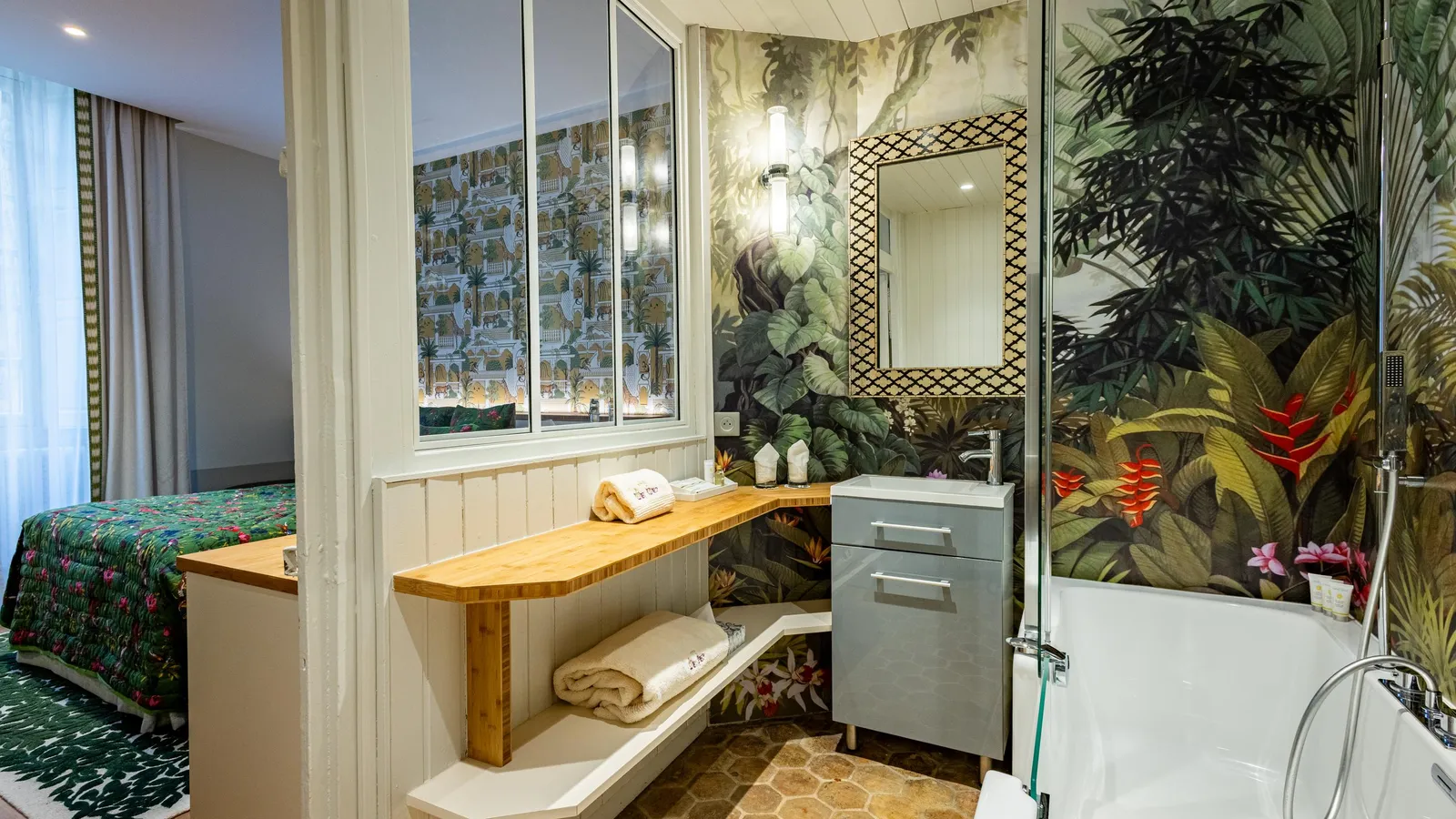 Salle de bain avec baignoire, lavabo et miroir, décorée de motifs végétaux, adjacente à une chambre avec lit et fenêtre