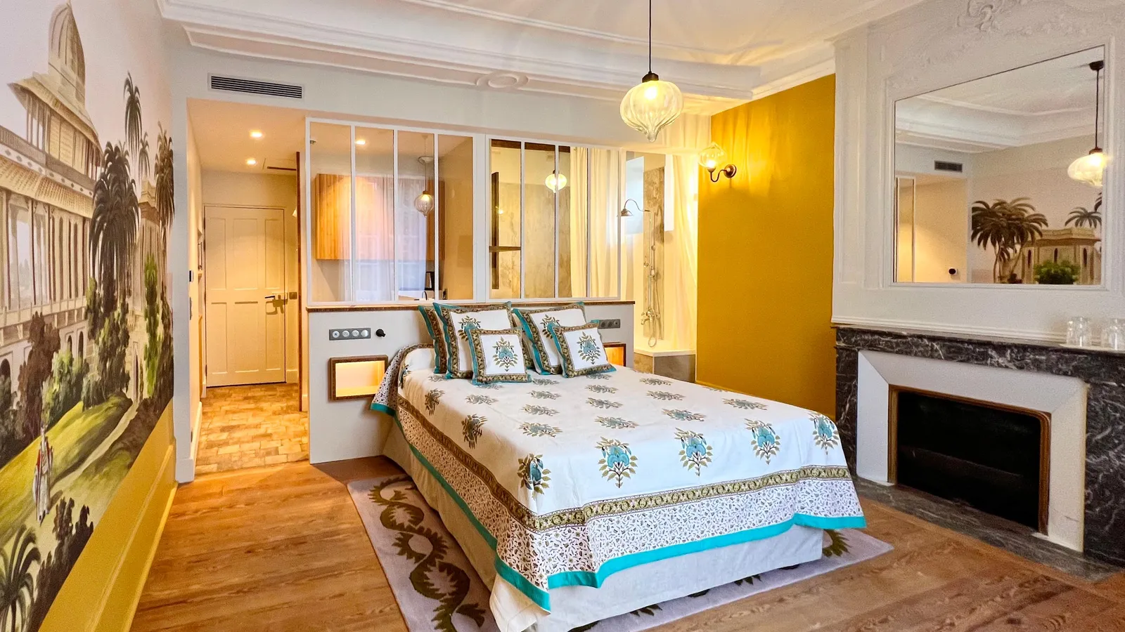 Chambre avec lit à baldaquin décoré de motifs floraux, cheminée en marbre, miroir et porte vitrée menant à une autre pièce