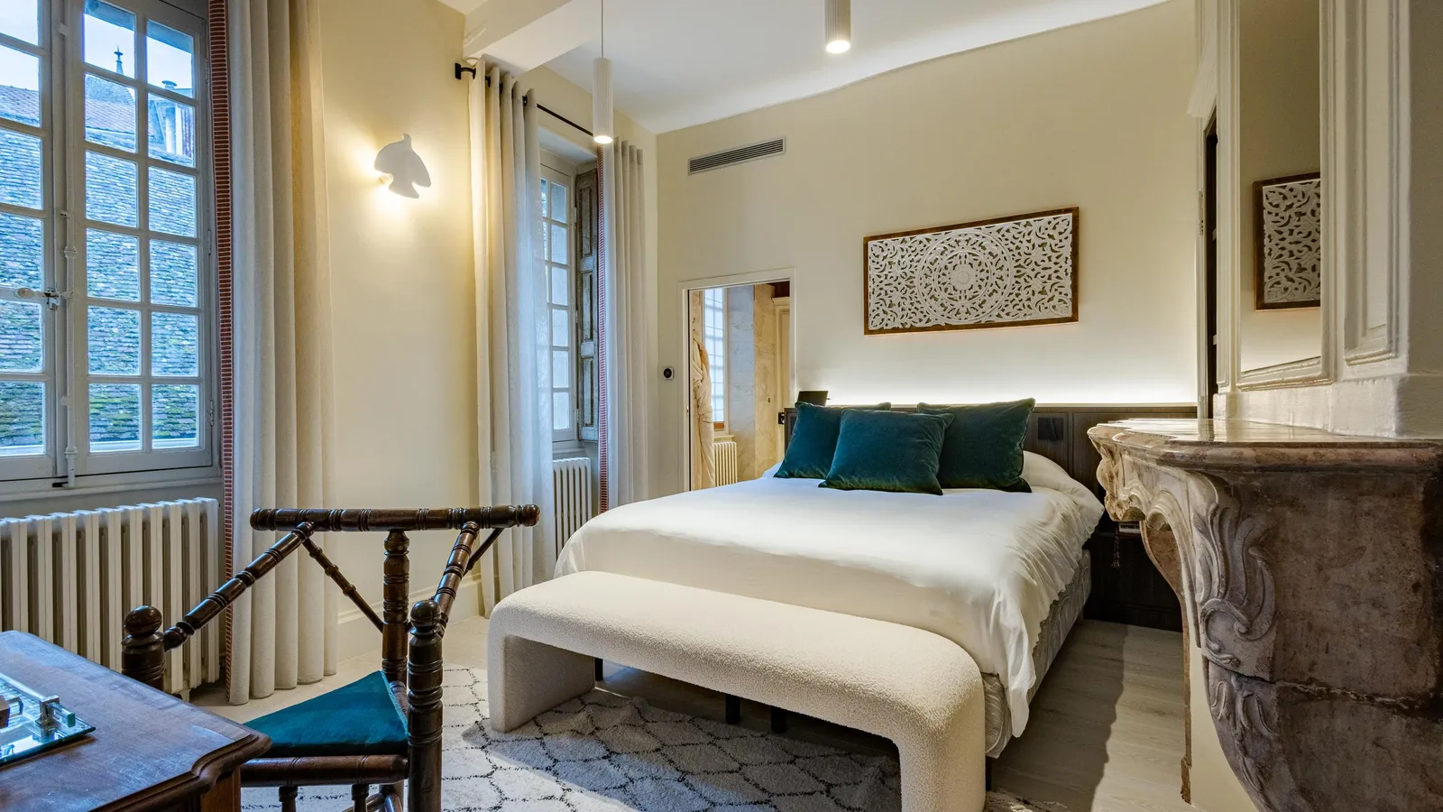 Chambre avec un lit double, une banquette au pied du lit, des fenêtres avec des rideaux et des radiateurs, décorée avec des coussins verts et des panneaux muraux