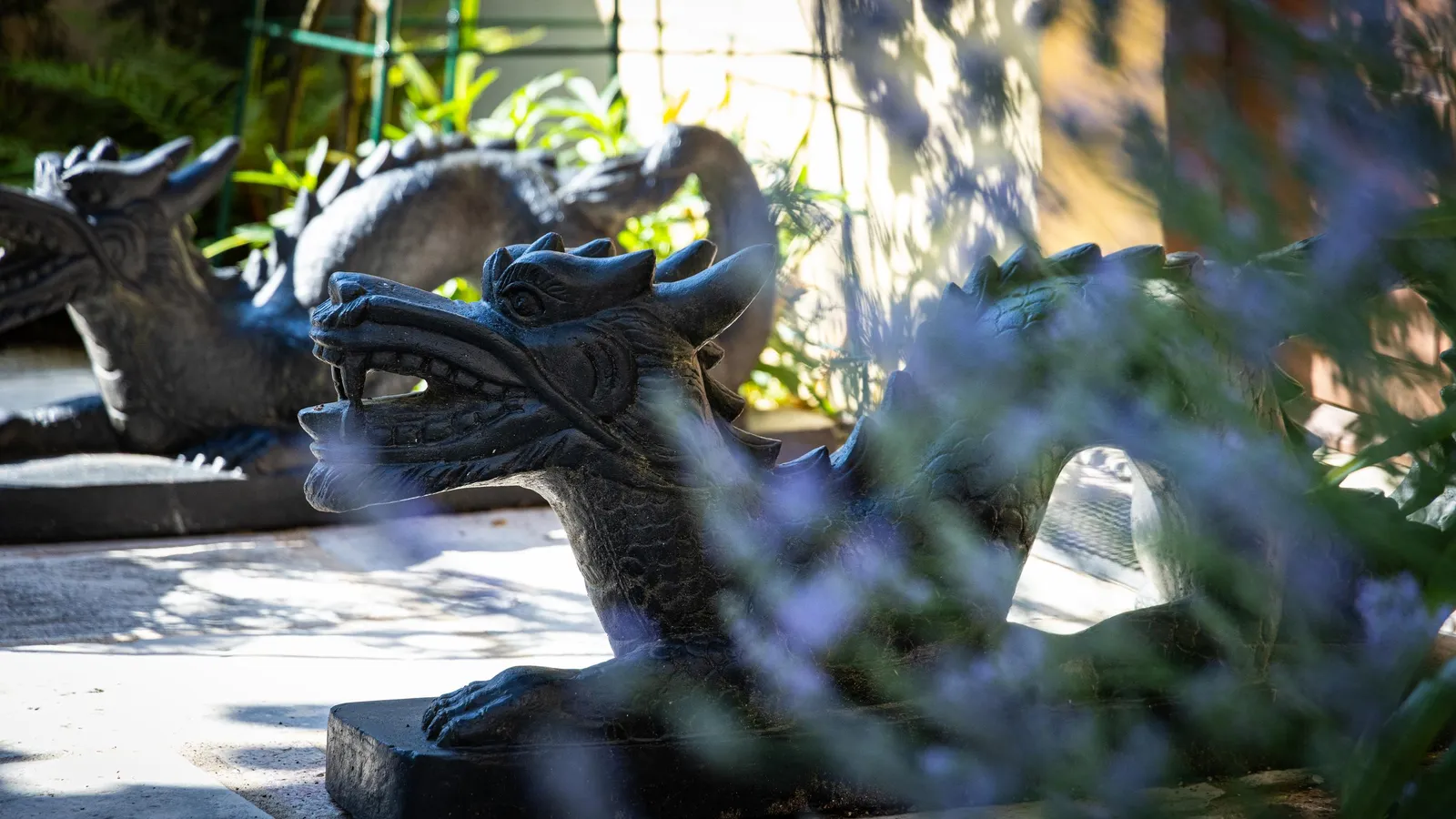 Statue de dragon en pierre noire posée sur un socle, dans un jardin avec des plantes en arrière-plan