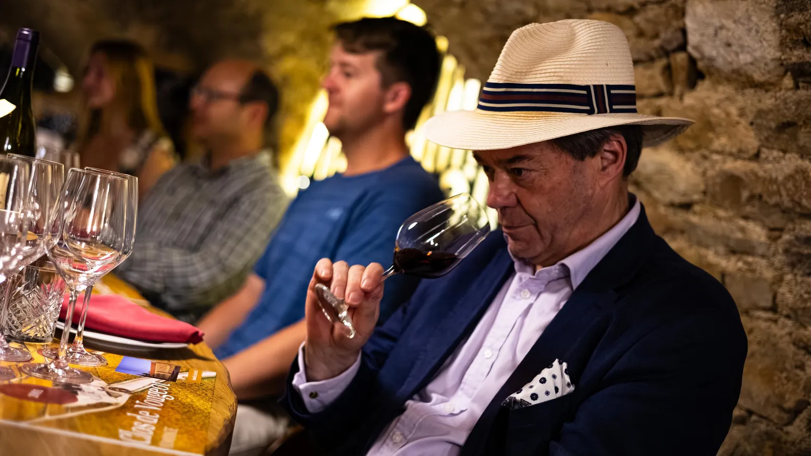 Un homme en costume et chapeau examine un verre de vin dans un cadre intérieur avec d'autres personnes en arrière-plan
