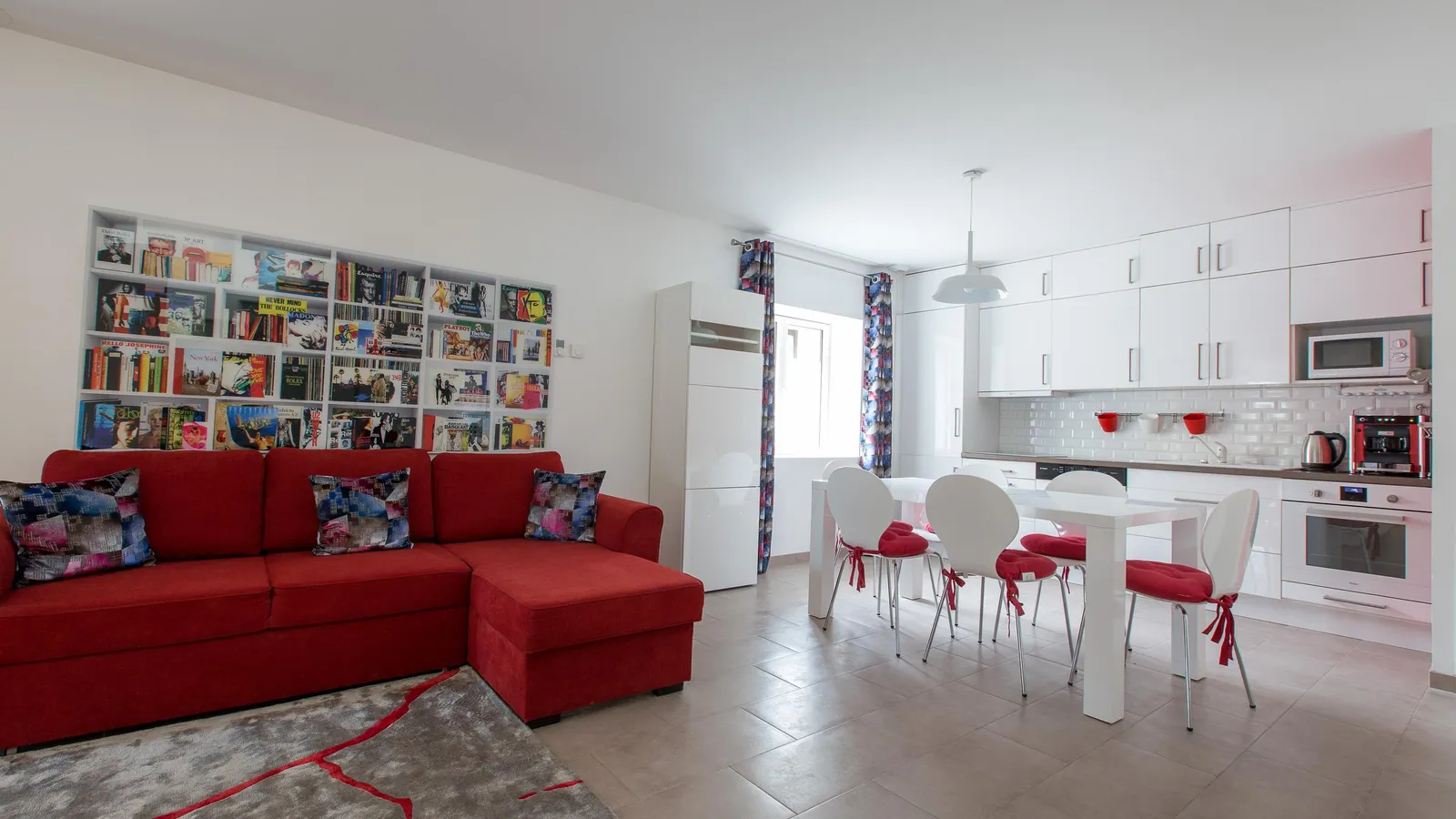 Living room with red sofa and book-filled library, white kitchen with dining table and red chairs