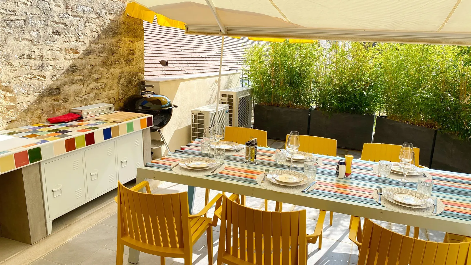 Table set outdoors under an awning with wooden chairs, a barbecue, and potted plants.