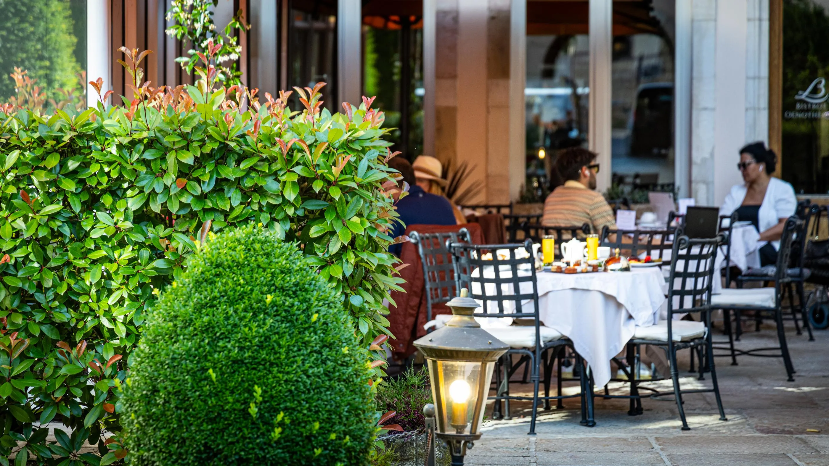 Des personnes sont assises à des tables en terrasse devant un restaurant, entourées de végétation et de lampadaires