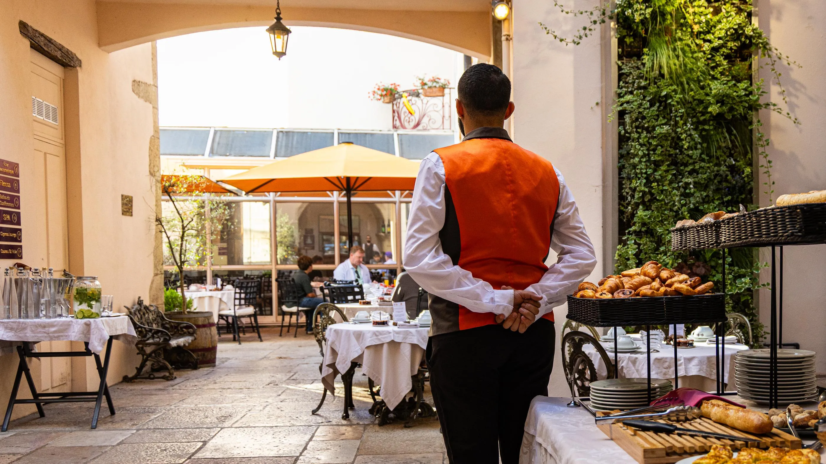 Un serveur en veste orange et chemise blanche se tient dos à la caméra dans un restaurant avec des tables dressées et des paniers de pain