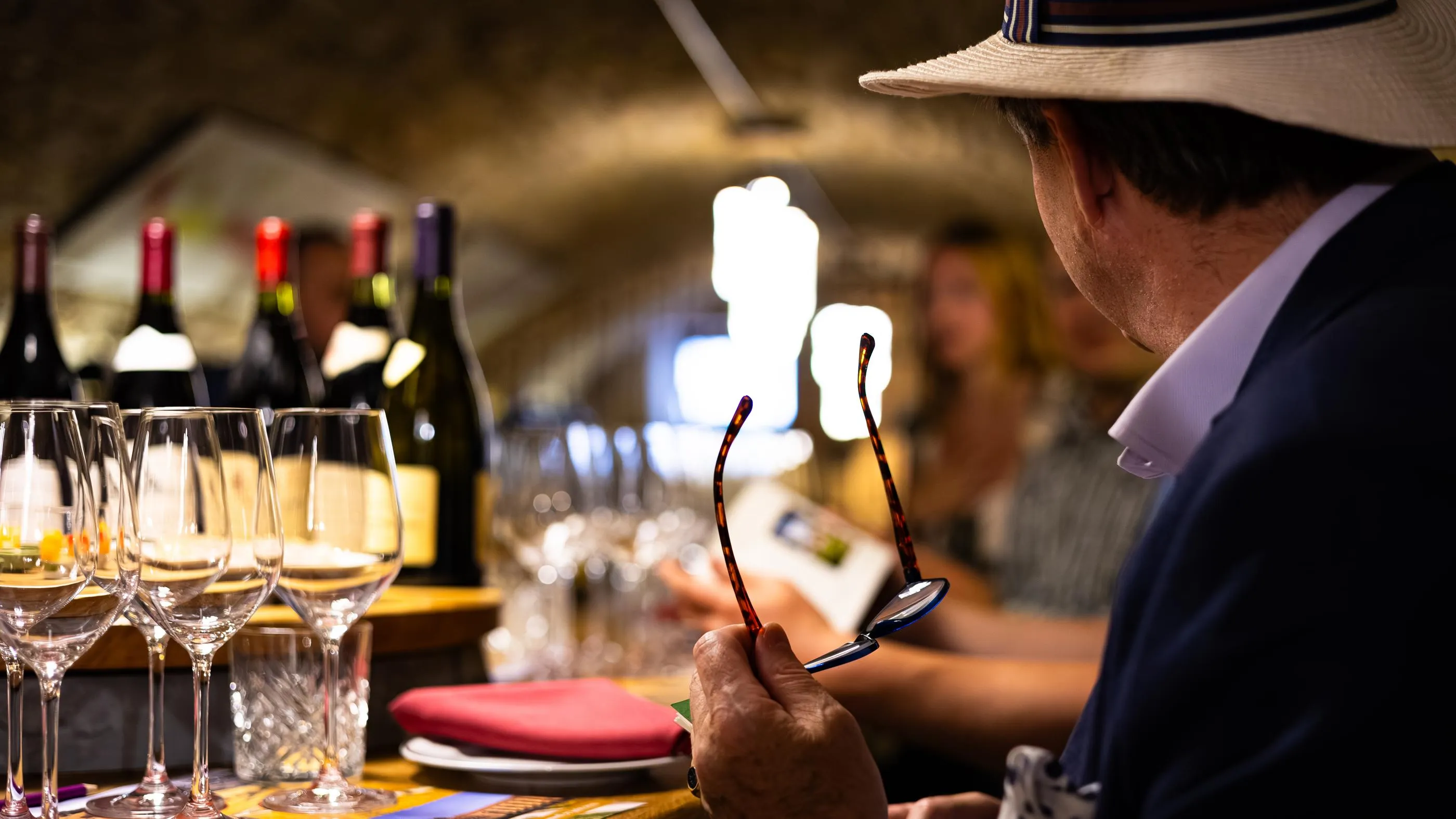 A man in a hat examines glasses in front of wine glasses and wine bottles.