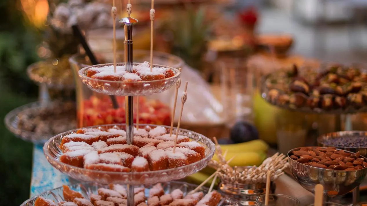 A three-tiered display of desserts with sugar-dusted pastries, bowls of nuts, and small pots of stuffed dates.
