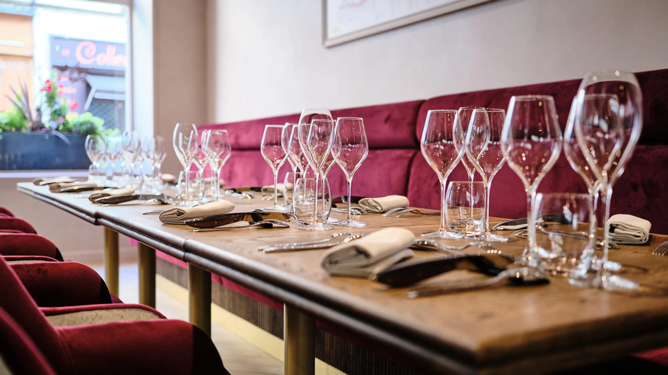 A restaurant table with wine glasses, cutlery, and napkins on a tablecloth, in front of a red bench.