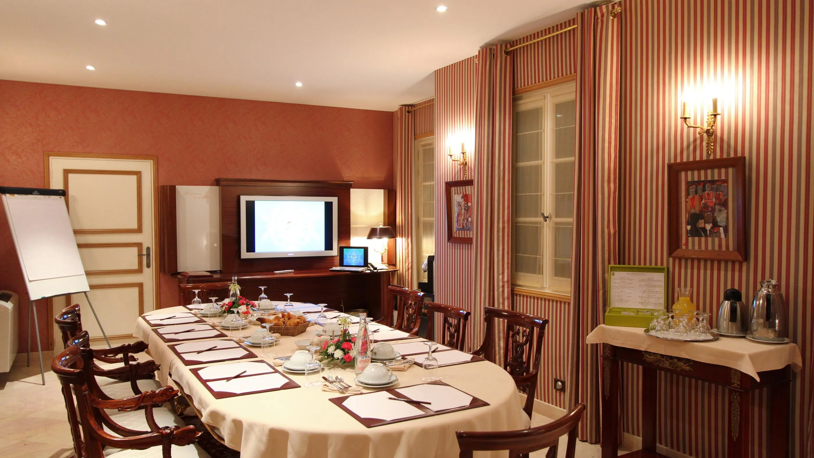 Meeting room with a rectangular table covered with a white tablecloth, wooden chairs, a screen, a whiteboard, and a service table with drinks.