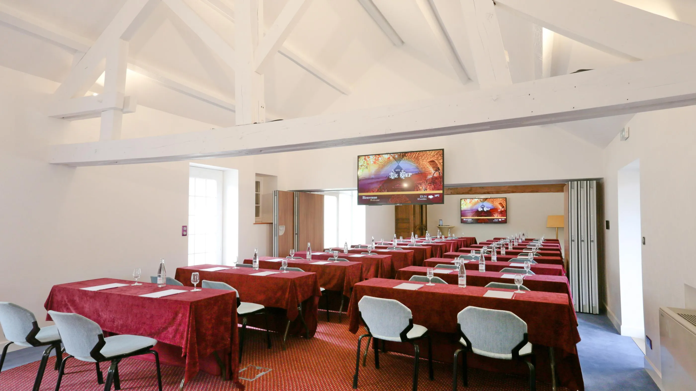 Meeting room with several tables covered with red tablecloths and chairs, equipped with two screens displaying the same content.