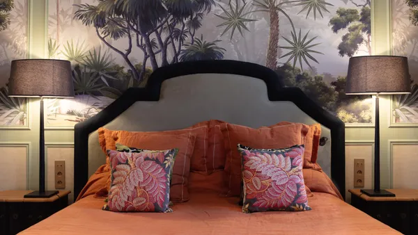 Bed with a gray headboard and orange and patterned cushions, flanked by two bedside lamps, in front of a wall decorated with palm patterns.