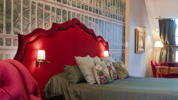 Chambre avec un lit à tête de lit rouge et une décoration murale représentant une bibliothèque, éclairée par des lampes murales et un lustre