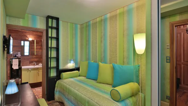Room with a bed covered by a blanket and green and blue cushions, a wall lamp turned on, a storage unit, and an open door leading to a wooden bathroom.