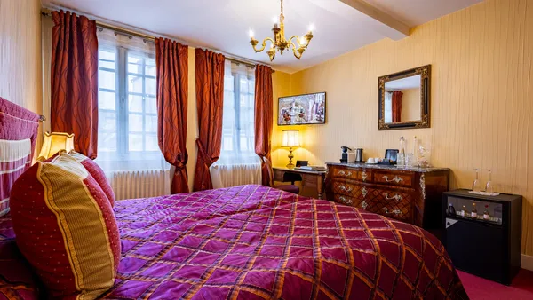 Chambre avec un grand lit à baldaquin recouvert d'une couverture à motifs rouges et or, des rideaux rouges aux fenêtres, un meuble de chevet avec une lampe allumée et un écran plat fixé au mur