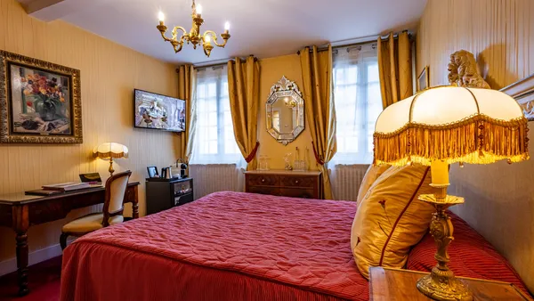 Room with a bed with a red cover, a bedside lamp, a desk, paintings on the walls, and a window with yellow curtains.