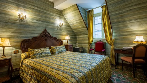 Bedroom with a four-poster bed, bedside lamps, a red armchair, and wooden walls with a window featuring yellow curtains.