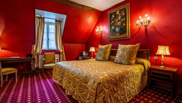 Bedroom with a four-poster bed covered in a golden blanket, bedside tables, a desk, and wall sconces; the walls are red and the floor is covered with a patterned rug.