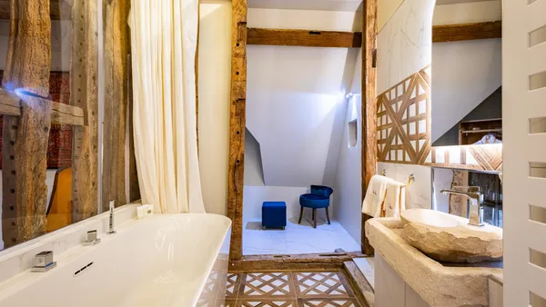 Baignoire blanche, lavabo en pierre et miroir dans une salle de bain avec des poutres en bois apparentes et une niche murale contenant deux tabourets bleus
