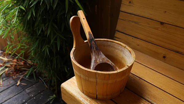 A wooden bucket with a ladle rests on a wooden stool near a wooden wall and green plants.