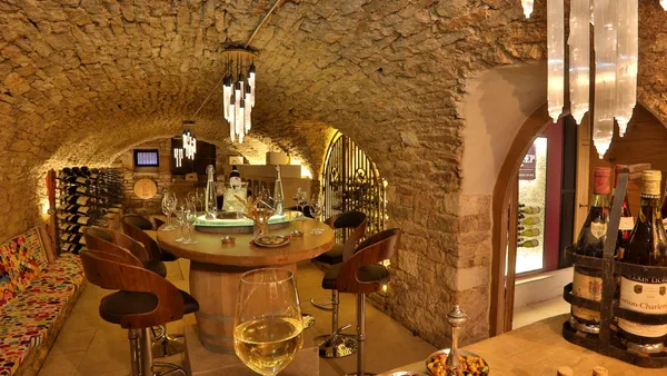 Un bar à vin souterrain avec des bouteilles de vin, des verres et des tabourets autour d'une table centrale, éclairé par des lumières modernes