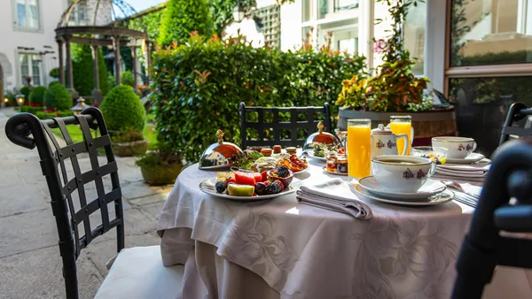 Table set outdoors with fruits, drinks, and dishes, surrounded by chairs and located in a garden with a pergola and vegetation.