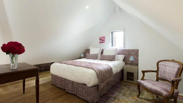 Room with a double bed covered by a patterned blanket, a bedside table with a lamp, a padded chair, and a table with a vase of red roses.