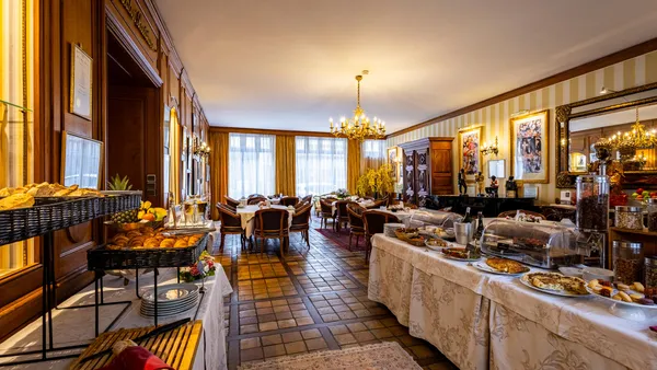 Elegant dining room with tables set with varied dishes, chandeliers, and large windows letting in natural light.