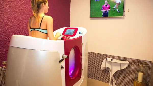 Une femme utilise un appareil de cryothérapie dans une pièce avec un écran affichant un match de rugby
