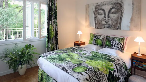Room with a double bed covered by a blanket featuring green and black leaf patterns, two bedside tables with lamps, a large wall painting, and windows overlooking a terrace surrounded by trees.