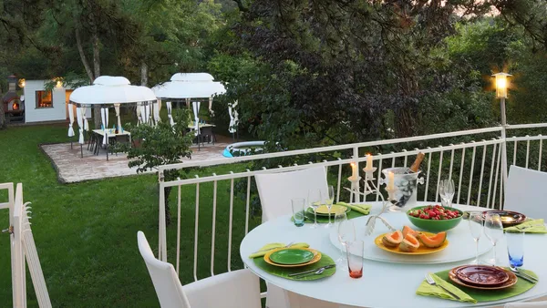Table set outdoors with plates, glasses, and candles, surrounded by white chairs, with umbrellas and a lawn in the background.