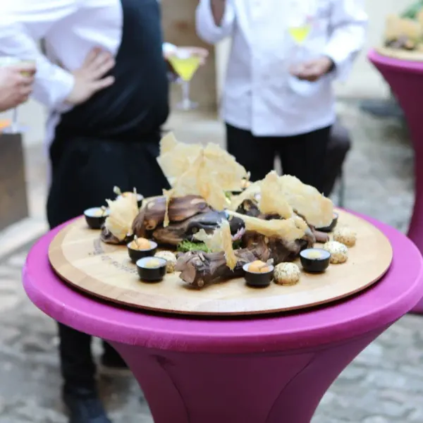 A dish of meat with garnishes and sauces is placed on a tall purple table, surrounded by people in formal attire holding glasses.